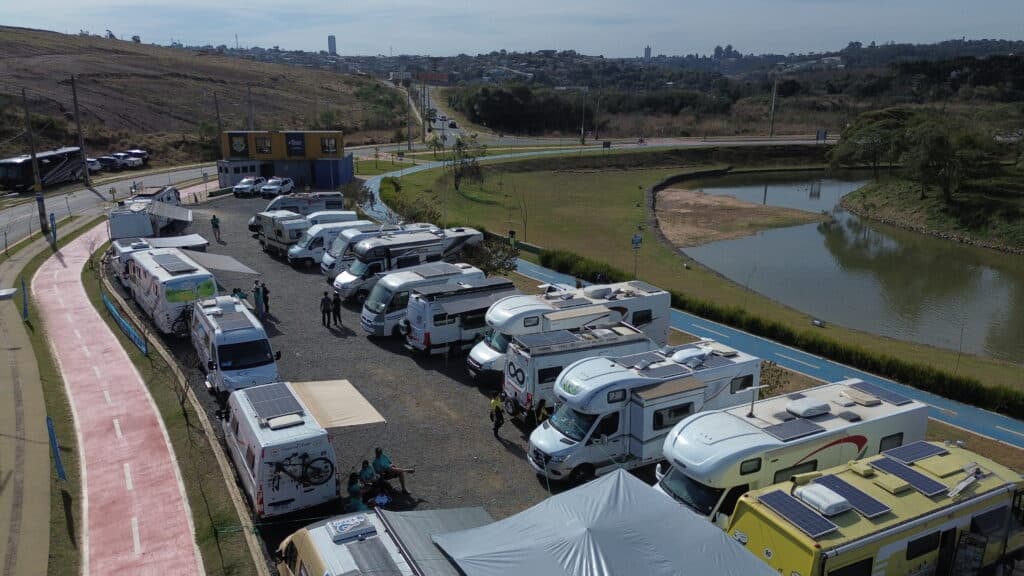 Na imagem, dezenas de motorhomes estão alinhados e estacionados ao redor do Lago de Olarias, em Ponta Grossa. O cenário ao fundo mostra o espelho d'água, áreas verdes e céu limpo.